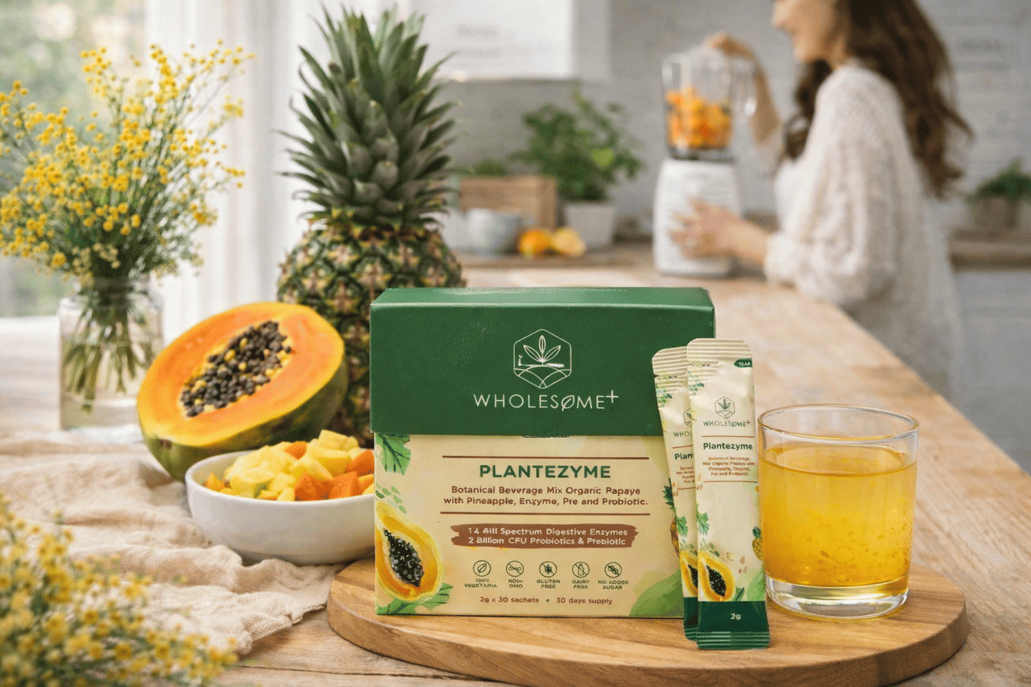 Wholesome+ Plantezyme box and sachets next to a glass of yellow drink on a wooden board, with papaya, pineapple, and a person blending fruit in a bright kitchen.