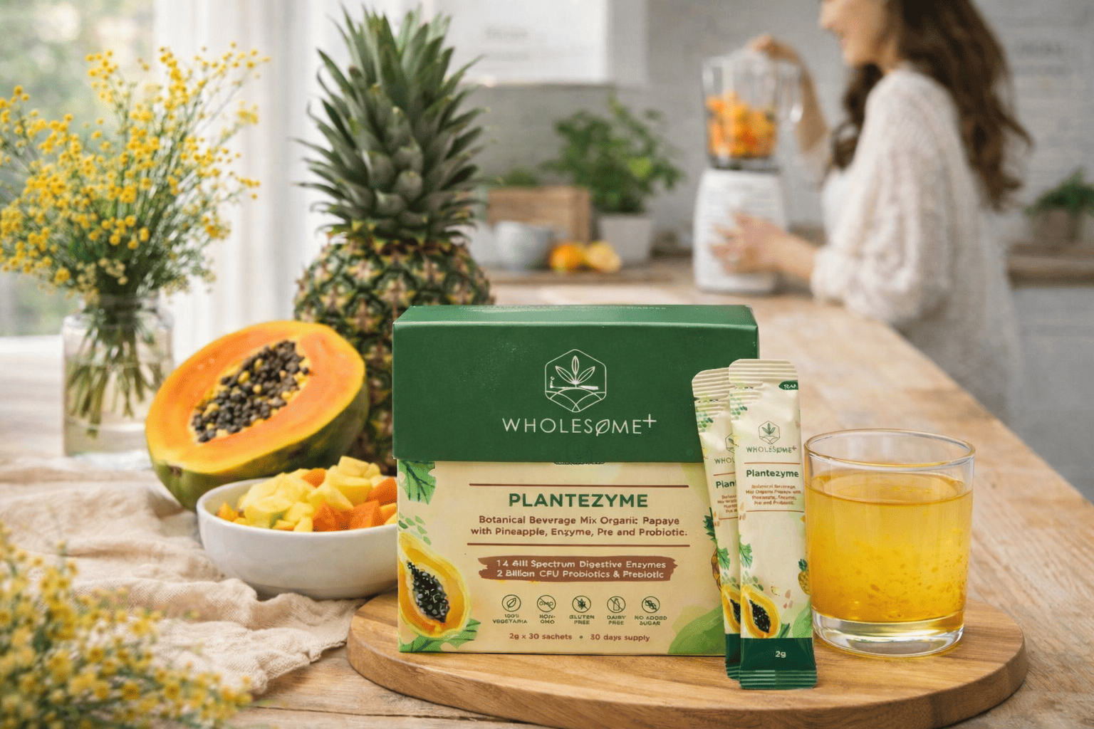 Wholesome+ Plantezyme box and sachets next to a glass of yellow drink on a wooden board, with papaya, pineapple, and a person blending fruit in a bright kitchen.